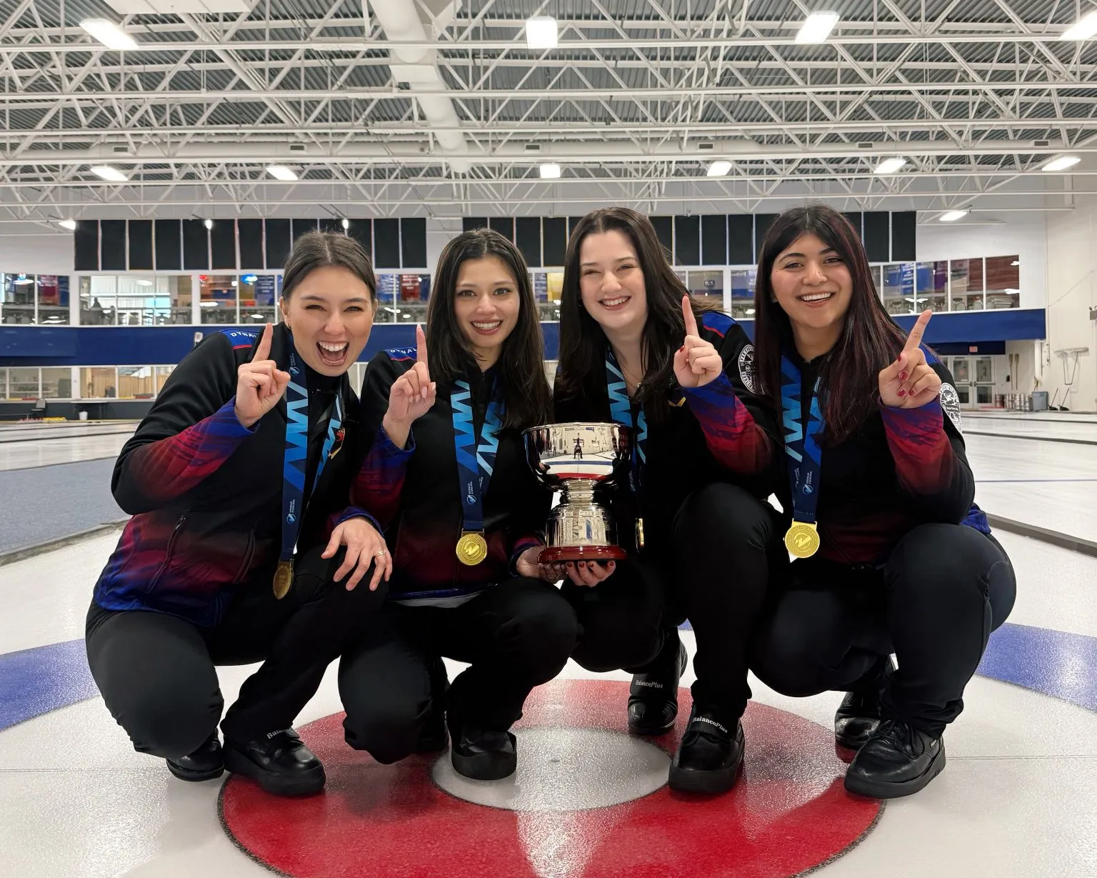 The four athletes celebrating with the trophy and gold medals, crouched on the curling house